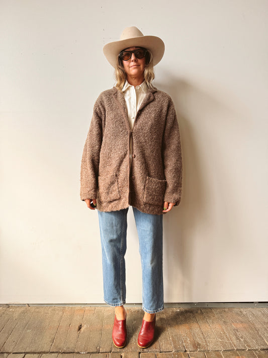 A woman stands against a plain off-white wall wearing a brown fuzzy button-front cardigan over a white shirt, blue jeans, and red boots, with a beige cowboy hat and dark sunglasses.