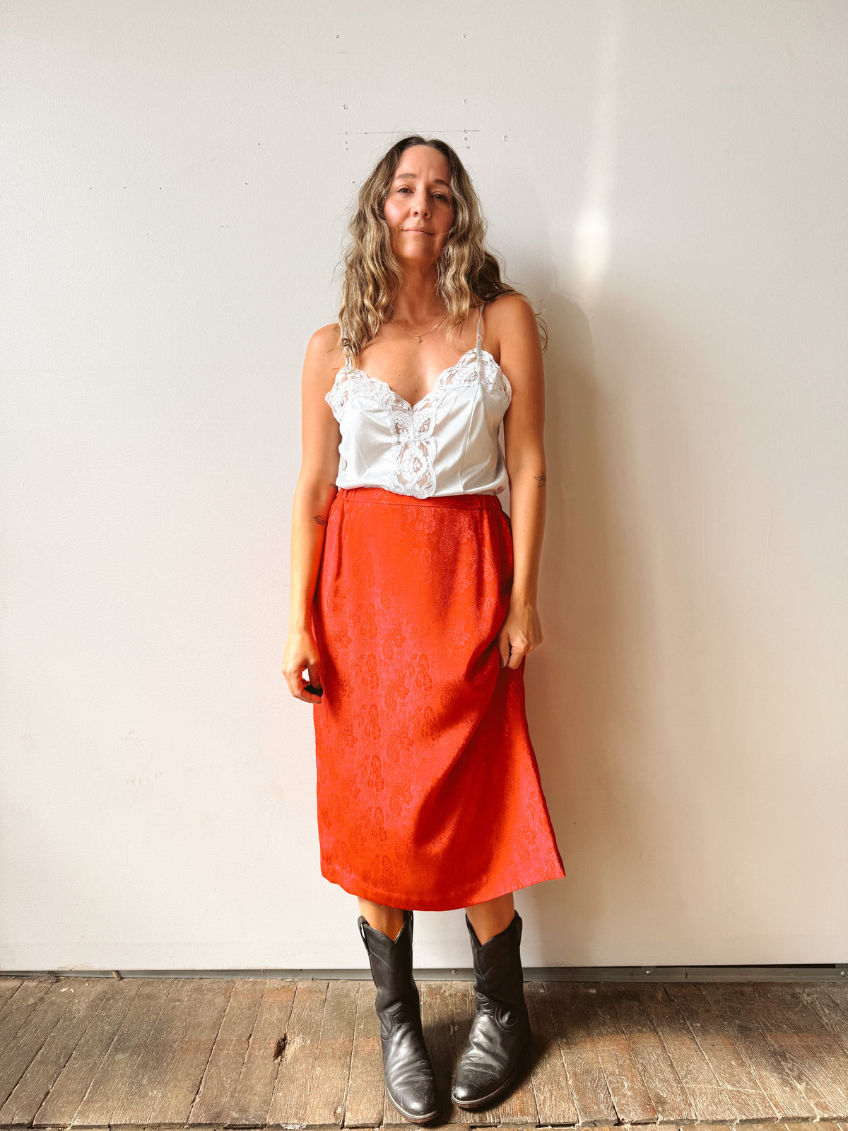 A woman stands against a plain off-white wall wearing a white lace-trimmed camisole top and a bright orange-red midi skirt, paired with black boots.