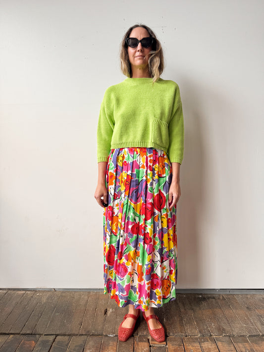 Person wearing a bright green sweater and colorful floral skirt against a plain background