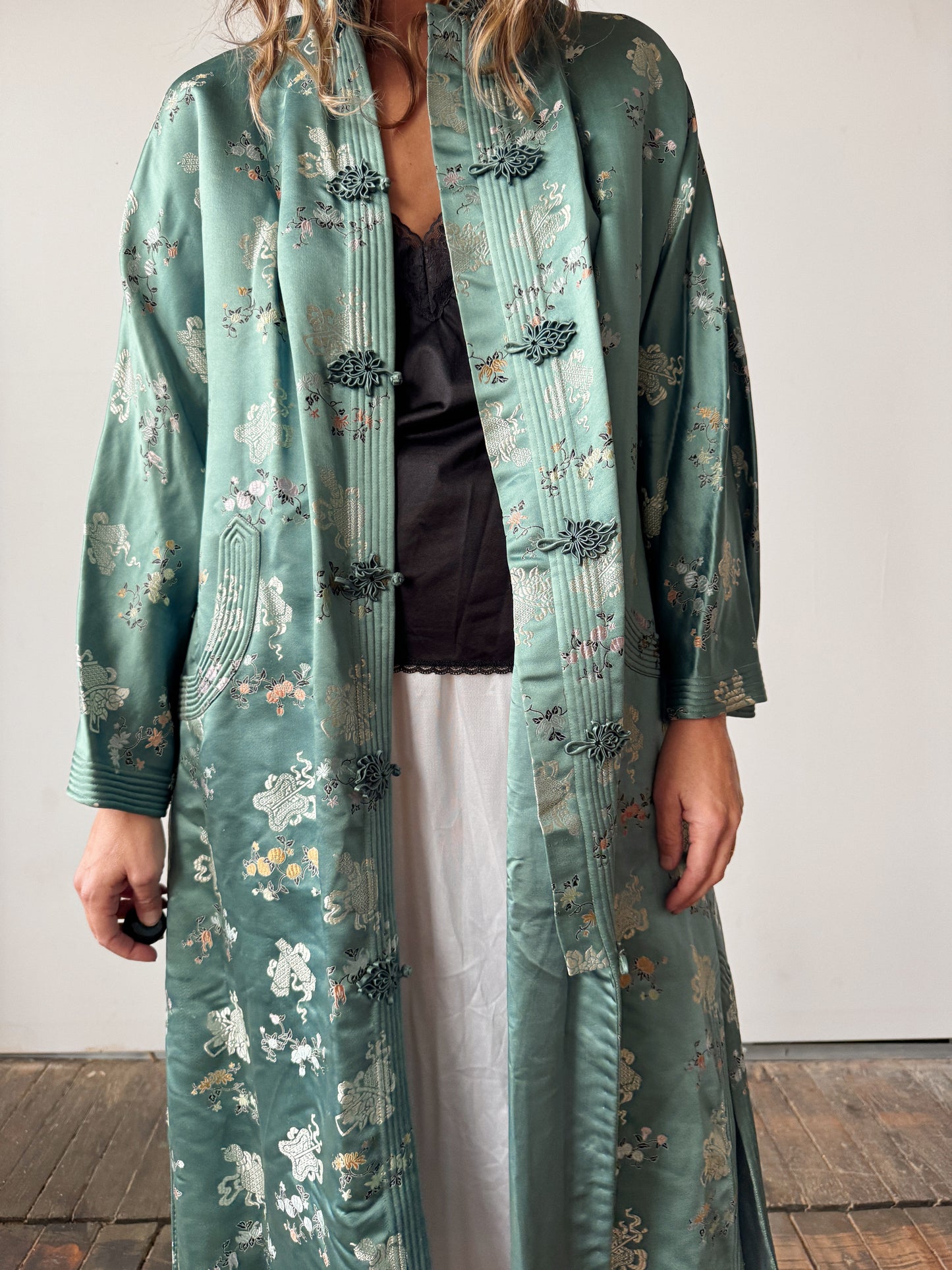 Person wearing a long, patterned green coat over a black top and white pants.