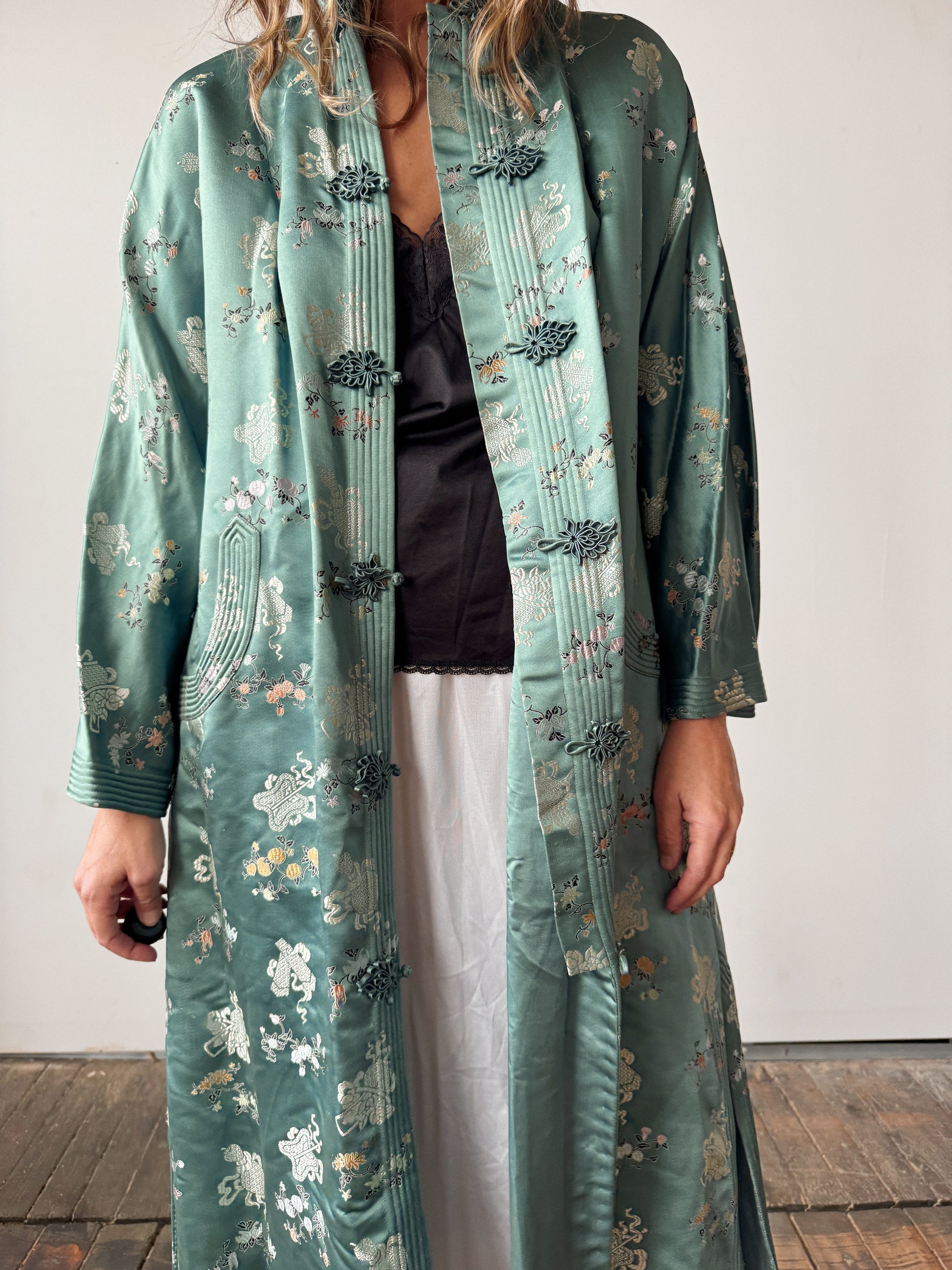Person wearing a long, patterned green coat over a black top and white pants.
